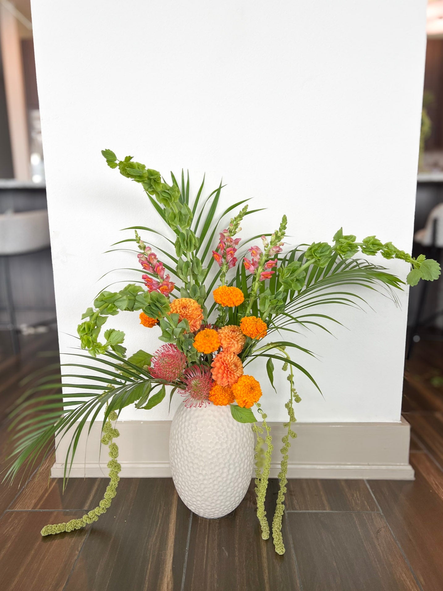 Plano floral arrangement with orange and pink flowers in a textured white vase on a wooden floor.
